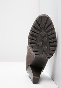 Bota de tacón alto de ante marrón con suela de goma texturizada que presenta un patrón de relieve y un tacón cuadrado. Vista lateral que muestra los detalles de la suela.