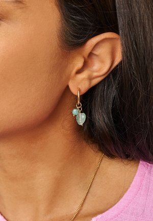 Orecchino a cerchio d'oro con due ciondoli di gemme verdi indossato sull'orecchio sinistro di una donna, che mostra capelli scuri e parte di una maglia rosa.