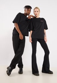 Two models wear matching black oversized t-shirts. One wears black flared pants; the other wears black trousers. Both have black shoes.