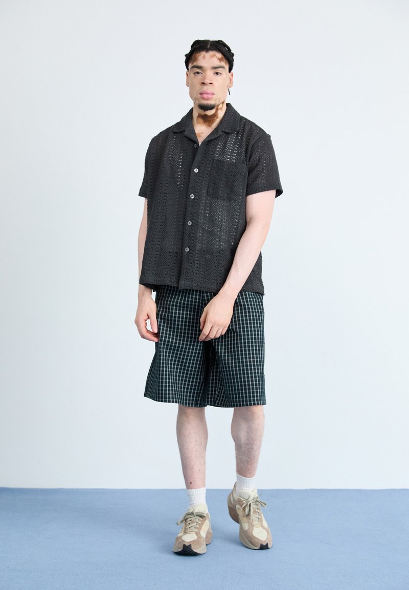 Maglietta nera a maniche corte e a maglia aperta con tasca, abbinata a pantaloni corti a quadri verdi. Il modello indossa sneakers beige e calzini bianchi.