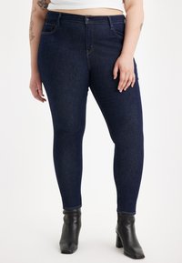 Jean ajustados de mezclilla en azul oscuro, con bolsillos frontales, costuras decorativas y una cintura alta. Combinados con botas negras de tobillo.