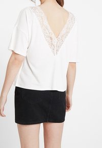 Bílý top s krátkým rukávem a V-výstřihem vzadu zdobený krajkou, kombinovaný s černou džínovou mini sukní se dvěma zadními kapsami.