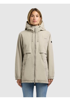 Frau trägt eine beige Kapuzenjacke mit Fronttaschen und Reißverschluss, steht vor einem einfarbigen grauen Hintergrund.