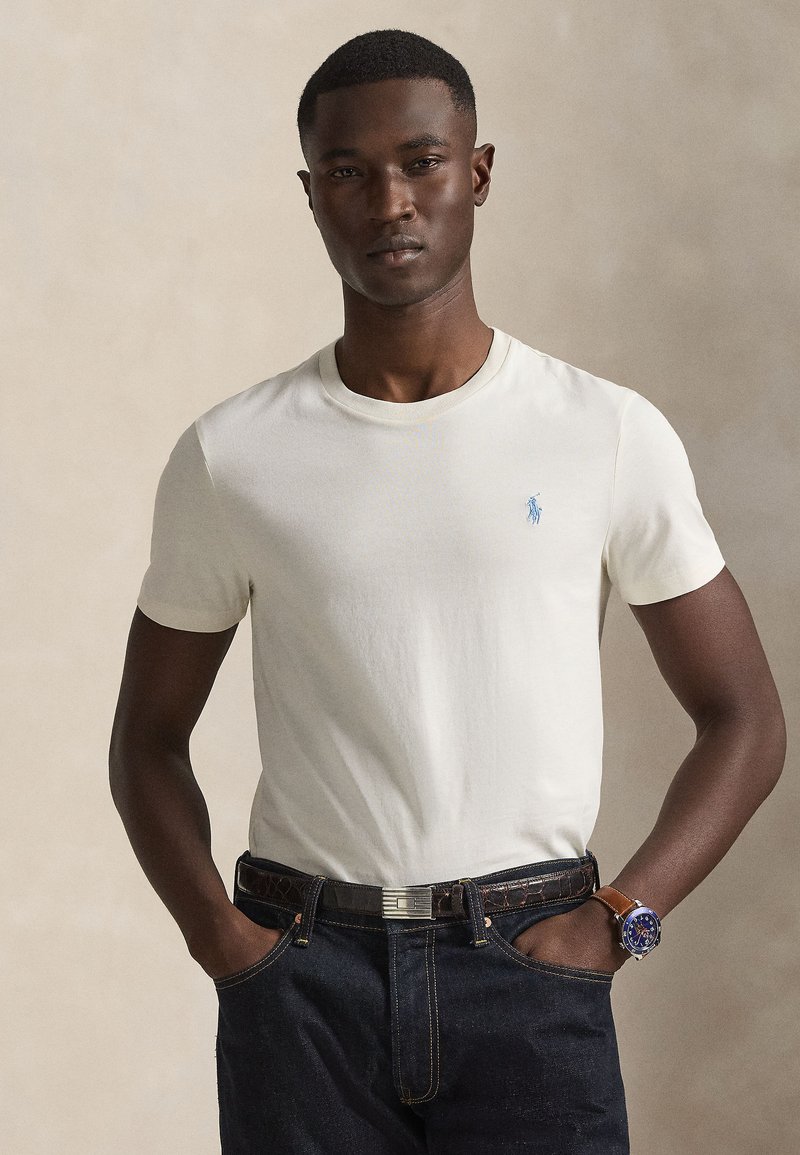 T-shirt blanc en coton avec un col rond et des manches courtes, comportant un petit logo bleu sur la poitrine, associé à un jean en denim foncé et une montre.