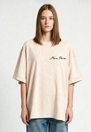 Jeune femme aux cheveux bruns raides porte un t-shirt beige ample avec le texte « Mira Paris » et un jean bleu, debout devant un fond uni.