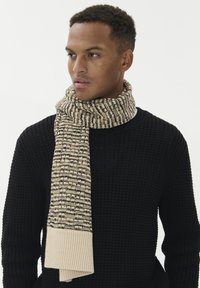 Strikket tørklæde i beige og brunt mønster med en ribbet lys beige ende. Bæres med en struktureret sort sweater med rund hals.