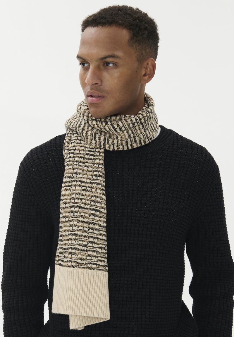 Strikket tørklæde i beige og brunt mønster med en ribbet lys beige ende. Bæres med en struktureret sort sweater med rund hals.