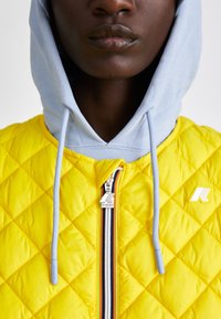 Veste matelassée jaune avec capuche bleue. Ses caractéristiques notables incluent une fermeture éclair à l'avant avec un tirant en métal, un tissu texturé et un accent logo.