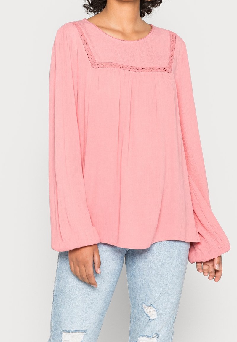 Femme portant un chemisier rose ample à manches longues avec bordure en dentelle au niveau du col et un jean bleu clair déchiré.