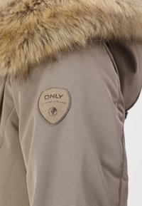 Parka ar bežīgu audumu, ar brūnu mākslīgās kažokādas apkakli un apaļu bežīgu nozīmīti ar tekstu "ONLY". Mīksta tekstūra un silts dizains.