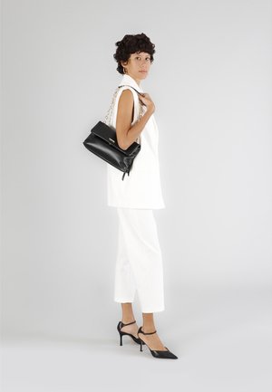 Borsa a mano nera in pelle con tracolla a catena, indossata sopra un blazer bianco senza maniche e pantaloni bianchi, completata da scarpe nere con tacco alto.