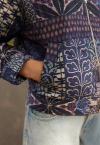 La chaqueta acolchada presenta una mezcla de patrones azules y morados, incluyendo diseños geométricos y florales. Cuenta con bolsillos y un cierre de cremallera.