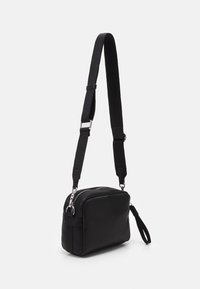 Bolso cruzado de cuero negro con correa de tela ajustable, forma rectangular, cierre con cremallera y asa lateral; textura suave y diseño minimalista.