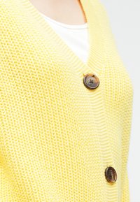 Κίτρινη πλεκτή ζακέτα με ντεκολτέ σε σχήμα V, δύο μεγάλα καφέ κουμπιά και υφή με ανάγλυφο σχέδιο. Ιδανική για να φορεθεί πάνω από διάφορα ρούχα.