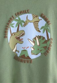 Grøn T-shirt med et cirkulært dinosaur-motiv med blå baggrund, palmetræer og teksten "DINOSAURS FAMILY RAWR!" i brun.