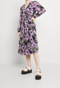 Květinové šaty s fialovými a růžovými květy na černém pozadí, s dlouhými rukávy, výstřihem do V a páskem v oblasti pasu. Nosené s platformovými sandály.