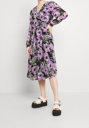 Robe florale avec des fleurs violettes et roses sur un fond noir, avec des manches longues, un col en V et une taille ceinturée. Portée avec des sandales à plateforme.