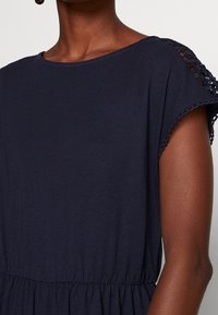 Granatowa sukienka z miękkiego materiału, z okrągłym dekoltem i koronkowymi detalami na ramieniu, z marszczoną talią podkreślającą kształty.