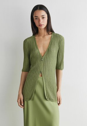 Vrouw met een geribd olijfgroen vest met twee knopen over een bijpassende olijfgroene rok, staand tegen een witte achtergrond.