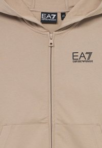 Beige zip-up hoodie made of soft fabric. Features an embroidered EA7 logo on the left chest and two front pockets. Smooth texture.
