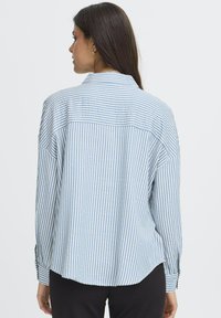 Woman with long dark hair wearing gold hoop earrings and a blue and white vertically striped long-sleeve shirt, facing away.