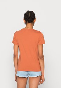 Persona con cabello trenzado vistiendo una camiseta de manga corta naranja y pantalones cortos de mezclilla azul claro, de pie de espaldas contra un fondo claro y liso.