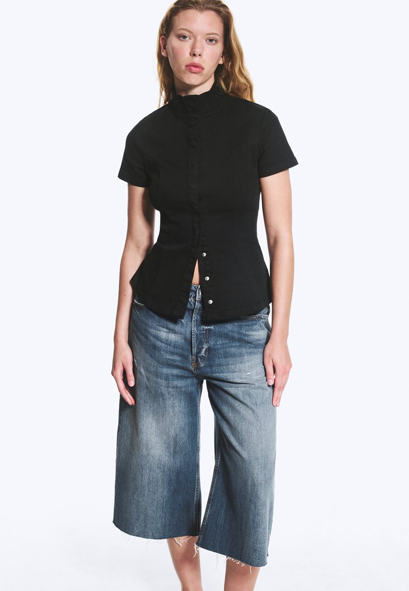 Femme portant une chemise noire à manches courtes boutonnée et un jean bleu en denim à jambes larges et coupé, sur fond blanc.