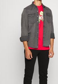 Muž v tmavě šedé džínové bundě s vyhrnutými rukávy, oblečený v červeném tričku s květinovým potiskem a černých kalhotách, stojící proti jednoduchému pozadí.