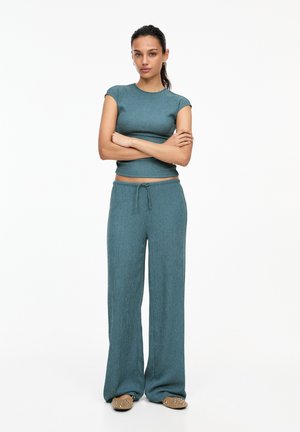 Top scurt tricotat ribbed în nuanță teal, asortat cu pantaloni largi, având un talie elastică și șnur. Modelul stă cu brațele încrucișate.