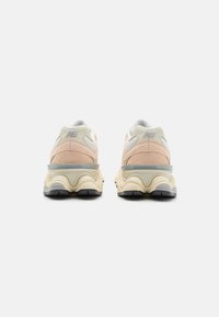 Kreemjas ja beežides New Balance'i sneakers'id, nähtavad tagant, koos massiivsete tallade ja võrku-kangast pealsetega valge tausta ees.