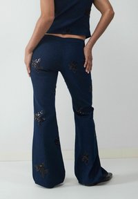 Marineblaue Schlaghosen mit floralen Paillettenverzierungen. Weicher Stoff, taillierte Passform und breite Säume. Kombiniert mit einem passenden Cropped Top.