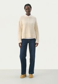 Suéter de punto de cable color crema con dobladillo acanalado, usado sobre una camisa blanca con cuello, combinado con jeans oscuros de pierna recta y zapatos de cuero deslizables color marrón.