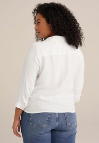 Camisa blanca de botones texturizada con cuello y mangas enrolladas; combinada con jeans de mezclilla azules. Vista desde la parte trasera, mostrando ajuste y estilo.