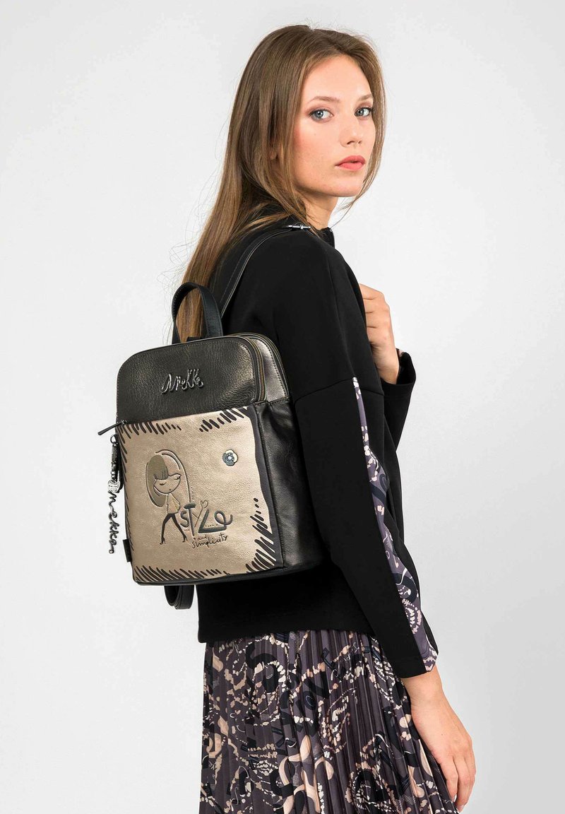 Mochila de cuero negro con una parte frontal de metal dorado, presenta un diseño de dibujos animados, cierre de cremallera y asa superior. Acabada con un charm.