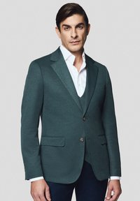 Ciemnoturkusowa marynarka wykonana z teksturowanej tkaniny, z pojedynczym zapięciem na guzik, klapami z wcięciem oraz dwoma przednimi kieszeniami. Ubrana na białą koszulę.