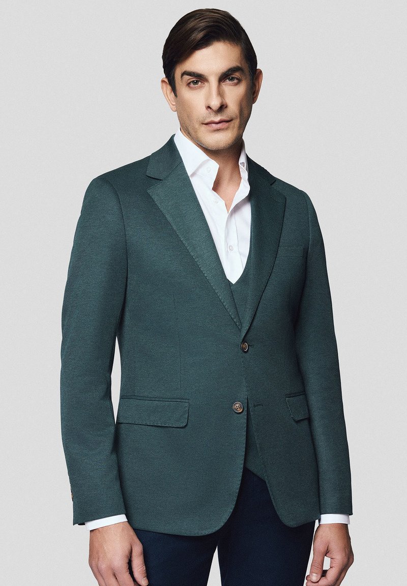 Ciemnoturkusowa marynarka wykonana z teksturowanej tkaniny, z pojedynczym zapięciem na guzik, klapami z wcięciem oraz dwoma przednimi kieszeniami. Ubrana na białą koszulę.