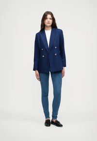 Marineblauwe dubbelzijdige blazer met gouden knopen, gecombineerd met een witte shirt en strakke blauwe jeans, aangevuld met zwarte instappers.