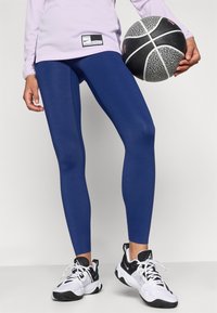 Zilās legingi no elastīga materiāla, kombinētā ar melniem un baltiem sporta apaviem. Kreisajā rokā turēts basketbola bumba, redzams lilac krāsas tops.