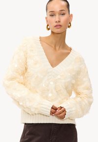 Pull crème avec un col en V, orné de pétales de tissu superposés et de sequins, présentant des bords côtelés pour une texture supplémentaire.