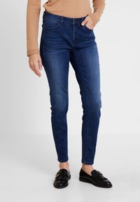 Persona vestida con jeans ajustados azul oscuro, camiseta de manga larga color beige y mocasines negros con detalle de flecos y hebilla dorada.