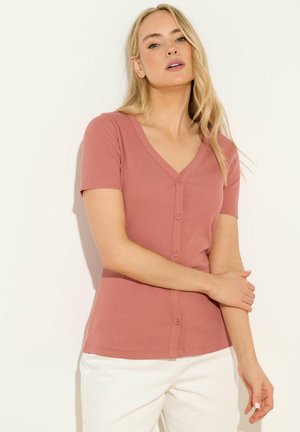 Blonde Frau trägt ein kurzärmliges, rosafarbenes Oberteil mit Knöpfen und weiße Hose, steht vor einem schlichten hellen Hintergrund.