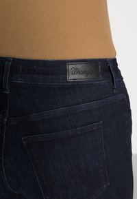 Jeans de mezclilla oscura con una textura suave, que cuentan con una etiqueta de marca de cuero negro adjunta a la cinturilla y un bolsillo trasero con detalle de costura.