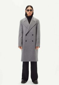 Grå ullblandning dubbelknäppt kappa med stora slag, svarta knappar, sidofickor, bärs över en svart polotröja och vida byxor.