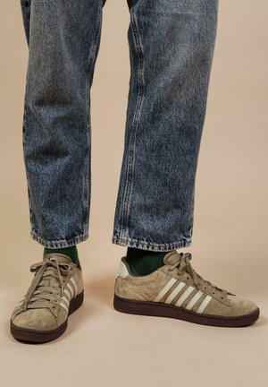 Beige suede sneakers with white stripes, featuring a lace-up design and brown rubber sole, paired with dark denim pants and green socks.