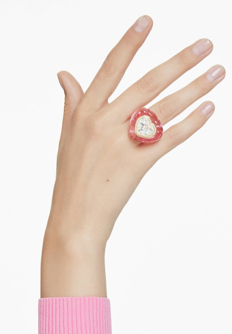 Hand med rosa tröja och hjärtformad rosa ring med stor klar ädelsten på långfingret mot vit bakgrund.