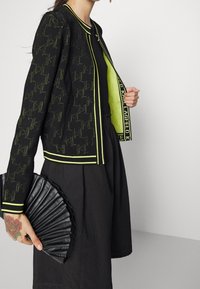 Czarna kurtka z wzorem pepitki, neonowo zielone akcenty i obszycia. Noszona na czarnym topie, z czarnym plisowanym wachlarzem w dłoni.