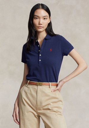 Vrouw met lang zwart haar draagt een marineblauw poloshirt met rood logo, beige hoge taille broek en een bruine riem, staat met de hand op de heup.
