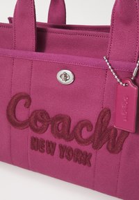 Sac en tissu violet Coach New York avec logo brodé, fermeture métallique à tourniquet et étiquette violette assortie sur chaîne à billes.