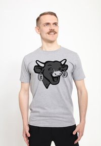 Graues Baumwoll-T-Shirt mit einem schwarzen, gepunkteten Kuh-Graphic, die Ohrringe und einen lächelnden Gesichtsausdruck zeigt. Kurzärmlig mit Rundhalsausschnitt.