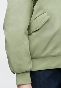 Ελαιόχρωμο bomber μπουφάν με ribbed στρίφωμα και μανσέτες, διαθέτει πλαϊνή τσέπη και ομαλή, ελαφρώς γυαλιστερή υφή υφάσματος.